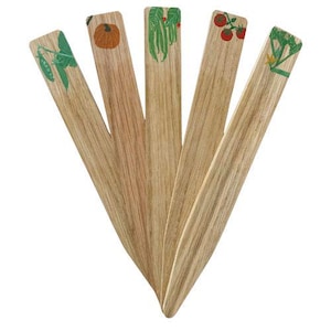 May include: Five wooden garden markers with painted illustrations of vegetables. The markers feature a pea plant, pumpkin, green beans, tomatoes, and a leafy plant. The markers are made of light-coloured wood.