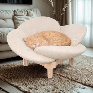 May include: A beige, flower-shaped cat bed with a sleeping orange tabby cat. The bed has a soft, textured fabric and wooden legs. The cat is curled up, appearing comfortable and relaxed.