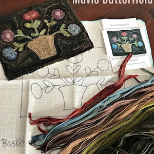 Op de afbeelding: Een close-up van een tapijt haakset met een gedrukt patroon voor een mand met bloemen. De set bevat een stuk canvas, garen en instructies. Het patroon is getiteld "A Basket of Blooming Posies" door Mavis Butterfield.