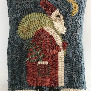 May include: A hand-stitched wool pillow featuring a whimsical depiction of Santa Claus carrying a sack of gifts and a Christmas tree. The pillow has a blue background with a white crescent moon in the upper right corner.