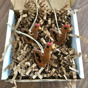 May include: Three brown ceramic chicken ornaments with red combs and black markings. The ornaments are hanging from twine and are in a box filled with brown packing paper.