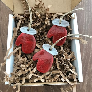 May include: Three red mitten ornaments with white tops and twine hangers are nestled in a cardboard box filled with brown paper filler. The ornaments are a festive holiday decoration.
