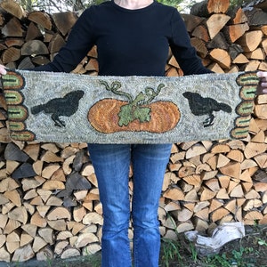 May include: A hand-stitched rug with a gray background, featuring two black birds and a large orange pumpkin with green leaves. The rug is held up by a person wearing blue jeans.