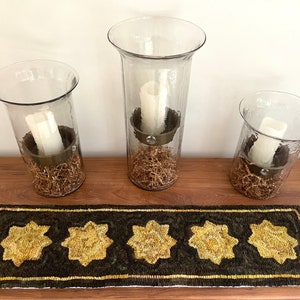 May include: Three clear glass candle holders of varying sizes, each with a white candle inside, sitting on a brown wooden surface with a gold and black patterned runner.