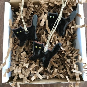 May include: Three black ceramic cat ornaments with yellow eyes, hanging from twine in a box filled with brown packing paper.