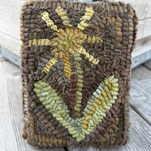 May include: Handmade, rectangular rug with a floral design. The rug features a central flower with yellow and green petals, set against a brown background. The rug is made of textured, looped yarn.