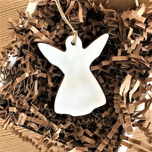 May include: White ceramic angel ornament with a string for hanging. The ornament is sitting in brown shredded paper.