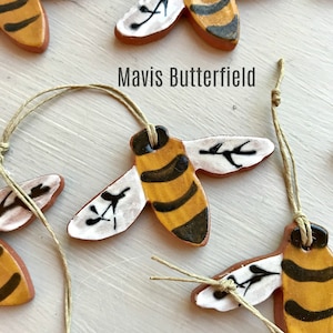 May include: A set of six hand-painted ceramic bee ornaments with brown, yellow, and black details. Each bee has a string attached for hanging. The ornaments are arranged on a white surface with the text "Mavis Butterfield" visible.