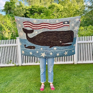 Op de afbeelding: Een rechthoekig haakwerk tapijt met een grote donkerbruine walvis en een kleinere walvis die in blauw water zwemmen. Boven de walvissen wappert een rood-wit-blauwe Amerikaanse vlag. Het ontwerp is versierd met witte sterren op een blauwe achtergrond.