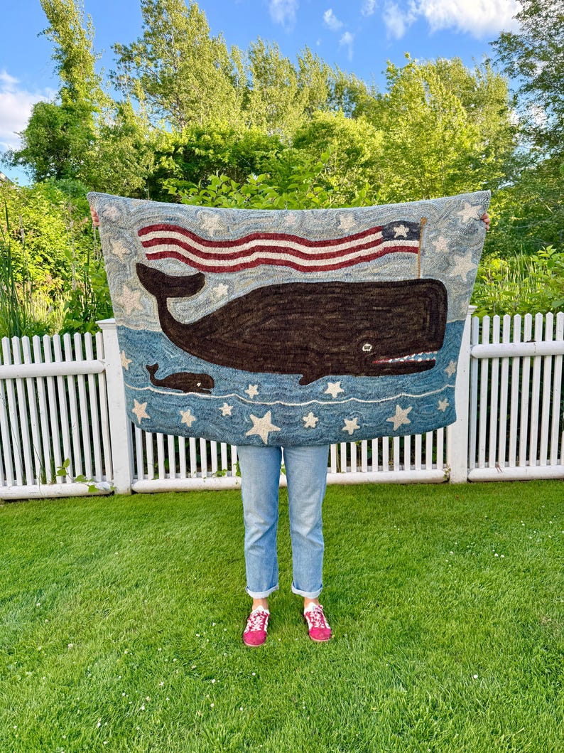 Op de afbeelding: Een rechthoekig haakwerk tapijt met een grote donkerbruine walvis en een kleinere walvis die in blauw water zwemmen. Boven de walvissen wappert een rood-wit-blauwe Amerikaanse vlag. Het ontwerp is versierd met witte sterren op een blauwe achtergrond.