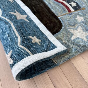 Op de afbeelding: Een rechthoekig gehaakt tapijt met een patriottisch ontwerp, voorzien van een donkerblauwe rand versierd met witte sterren, een effen donkerbruine middenbaan en een sectie met rode en witte strepen en een blauw veld met witte sterren.