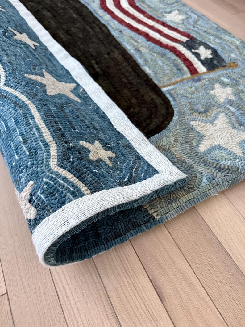 Op de afbeelding: Een rechthoekig gehaakt tapijt met een patriottisch ontwerp, voorzien van een donkerblauwe rand versierd met witte sterren, een effen donkerbruine middenbaan en een sectie met rode en witte strepen en een blauw veld met witte sterren.