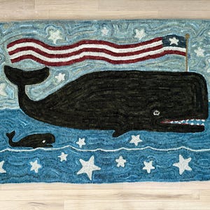 May include: A rectangular hooked rug featuring a large black whale and a smaller black whale swimming in a blue sea filled with white stars. Above the large whale, a red, white, and blue American flag waves against a light blue starry sky.