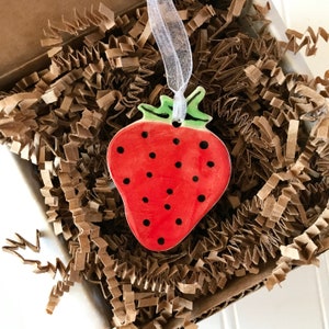 May include: A red and green ceramic strawberry ornament with black seeds. The ornament is hanging from a white ribbon.