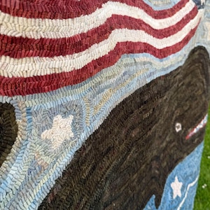 Op de afbeelding: Een kleed met een grote donkerbruine walvis die door een lichtblauwe zee vol witte sterren zwemt. Boven de walvis strekt zich een golvend patroon van de Amerikaanse vlag in rood, wit en blauw uit over het bovenste deel van het textiel.