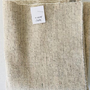 May include: Folded beige fabric with a textured weave and small flecks of color. A small white tag is attached with the handwritten words "morse code". The fabric has a raw edge.