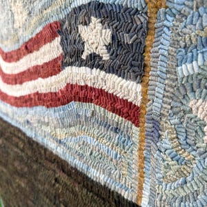 Op de afbeelding: Een handgehaakt kleed met een textuur van rode, witte en blauwe garenlussen in de vorm van een Amerikaanse vlag. Het ontwerp toont een witte ster op een donkerblauw veld, rode en witte strepen en een goudbruine vlaggenmast tegen een blauwe achtergrond.