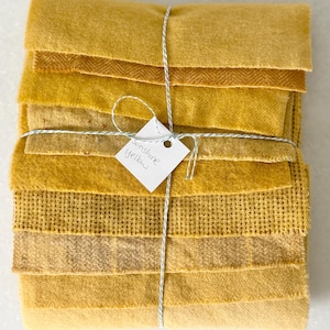 May include: A stack of fabric swatches in various shades of yellow, tied together with white and grey twine. A small white square tag is attached to the twine with the handwritten text Sunshine yellow. The fabrics feature different textures.