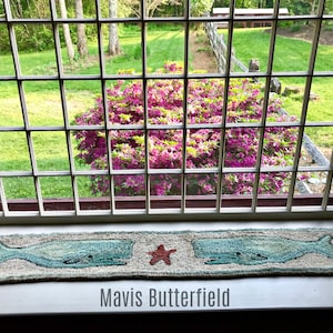 May include: A window with a view of a green lawn and a bush of pink flowers. A rug with two blue whales and a red starfish is on the windowsill. The text "Mavis Butterfield" is below the rug.