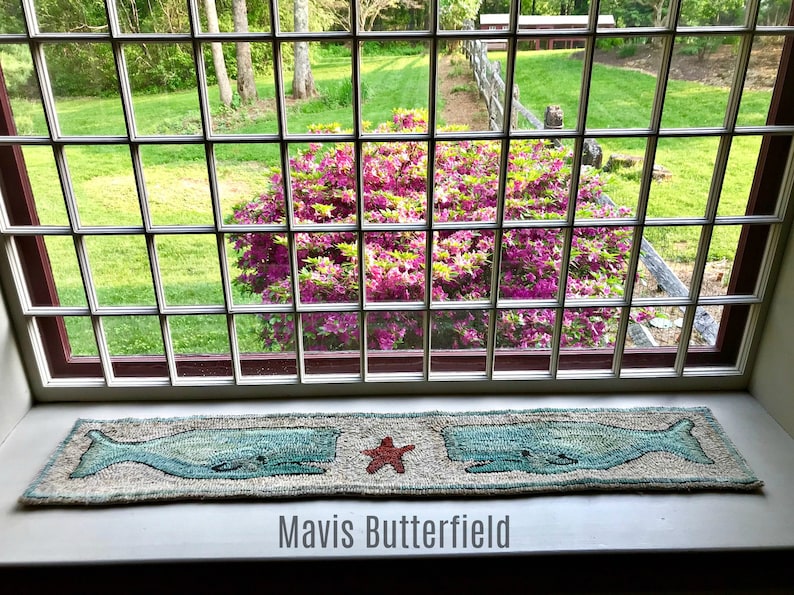 May include: A window with a view of a green lawn and a bush of pink flowers. A rug with two blue whales and a red starfish is on the windowsill. The text "Mavis Butterfield" is below the rug.