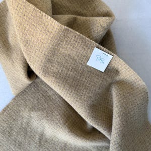 May include: A folded, tan-colored scarf with a textured weave. The scarf is made of a soft material and has a label that reads "Poffie". The scarf is draped, showing its length and texture.