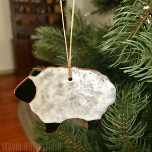 May include: A ceramic sheep ornament with a white body, black head and legs, and a brown rim. The ornament is hanging from a natural twine string, and is displayed on a Christmas tree. The ornament is handmade.