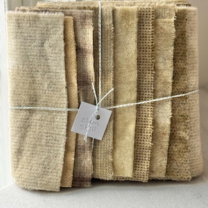 May include: A bundle of fabric squares in various shades of beige and brown, tied with white string. A small tag with the text "Clam Shell" is attached. The fabrics have different textures, including woven and textured surfaces, suggesting a craft or quilting project.