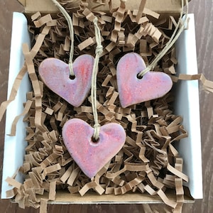 May include: Three heart-shaped ceramic ornaments with a pink and orange glaze, each strung on a natural twine cord. The ornaments rest in a white box filled with brown paper packing material. The hearts have a hole at the top for hanging.