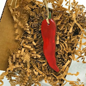 May include: A red chili pepper ornament with a green stem, hanging from a twine string in a box filled with brown packing paper.