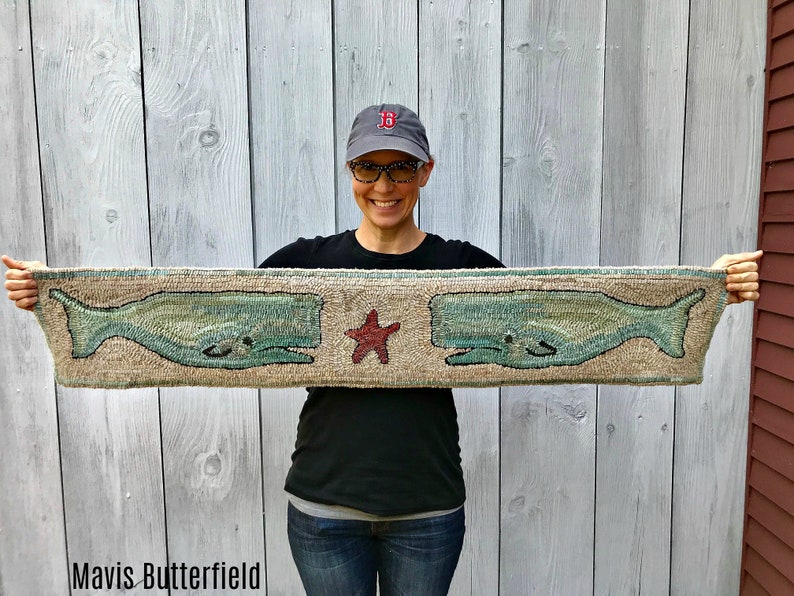 May include: A woman is holding up a beige rug with two green whales and a red starfish embroidered on it. The rug is approximately 45 cm long and 15 cm wide.  "Mavis Butterfield" is written in black text at the bottom of the rug.
