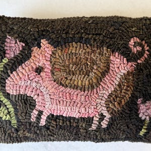 May include: A pink and brown hooked rug depicting a pig with flowers on either side. The rug is made of wool and has a textured surface.