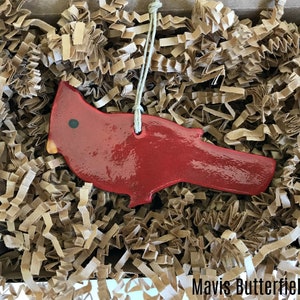 May include: A red cardinal bird ornament made of ceramic with a string for hanging. The ornament is sitting in a box filled with brown packing paper.