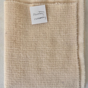 May include: Folded beige fabric with a textured surface. A small white tag with the handwritten words "country creamery" is pinned to the fabric. The fabric has a soft, neutral color.