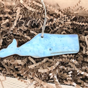 May include: A light blue ceramic whale ornament with a speckled glaze. The ornament is hanging from a brown string and is sitting on a bed of brown packing paper.