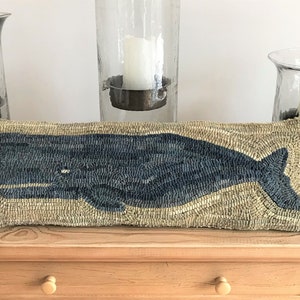 May include: A blue whale embroidered on a tan pillow. The pillow is on a wooden table with two glass candle holders on either side.
