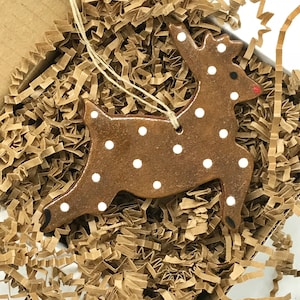 May include: A brown ceramic reindeer ornament with white polka dots. The ornament is hanging from a string and is in a box filled with brown packing paper.