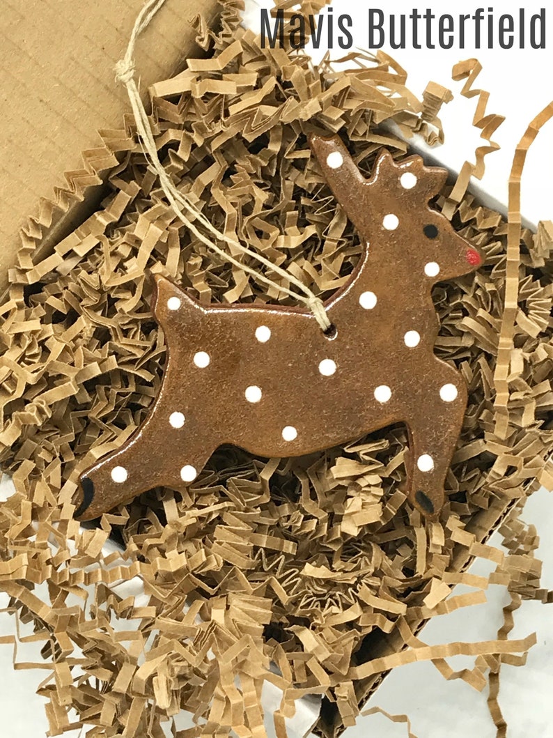 May include: A brown ceramic reindeer ornament with white polka dots. The ornament is hanging from a string and is in a box filled with brown packing paper.