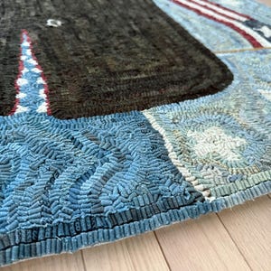 Op de afbeelding: Een rechthoekig kleed met een grote donkerbruine walvis in het midden op een lichtblauwe achtergrond met gestructureerde golfpatronen. Het ontwerp bevat een rood-wit gestreept randelement en kleine witte stermotieven, gemaakt met een lusjes-techniek.
