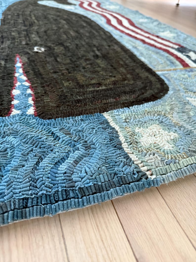 Op de afbeelding: Een rechthoekig kleed met een grote donkerbruine walvis in het midden op een lichtblauwe achtergrond met gestructureerde golfpatronen. Het ontwerp bevat een rood-wit gestreept randelement en kleine witte stermotieven, gemaakt met een lusjes-techniek.