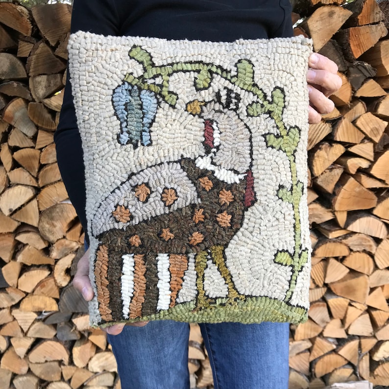 May include: A hand-stitched rug hooking pillow featuring a brown and white turkey with a green leafy vine and a blue flower. The pillow is on a beige background.