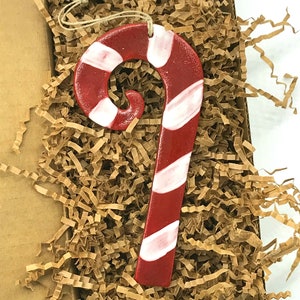 May include: A red and white candy cane Christmas ornament in a brown paper gift box with shredded paper. Mavis Butterfield is written on the bottom of the box.