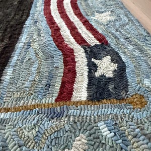 Op de afbeelding: Een handgeknoopt tapijt met een textuurontwerp van een wapperende Amerikaanse vlag met rode, witte en blauwe strepen en een donkerblauw veld met witte sterren. Het tapijt bevat een horizontale goudkleurige stok en een grote witte ster onderaan.
