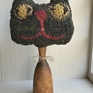 May include: A handcrafted cat-shaped decorative item. The cat's face is made of dark green and black textured fabric, with yellow and tan eyes and a red nose and mouth. It is mounted on a wooden base with a twine bow.