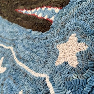Op de afbeelding: Een kleed met een donkerbruine walvis met een rood-wit patroon in de bek, zwemmend door een getextureerde blauwe zee. Het ontwerp bevat witte sterren en een golvende witte lijn, gemaakt met een dichte lus-techniek op een stoffen basis.