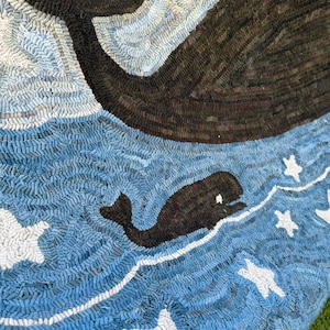 Op de afbeelding: Een kleed met een grote donkerbruine walvisstaart aan de bovenkant en een kleinere donkerbruine walvis die in een blauwe oceaan zwemt. Het ontwerp bevat witte sterren verspreid over het blauwe water, gemaakt met een getextureerde luspooltechniek.