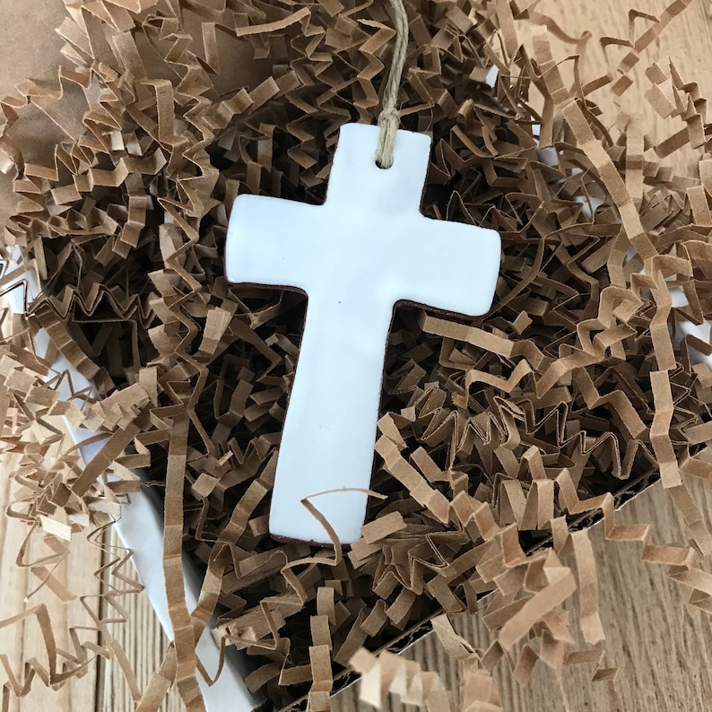 May include: White ceramic cross ornament with a twine hanger. The cross is sitting in brown shredded paper.