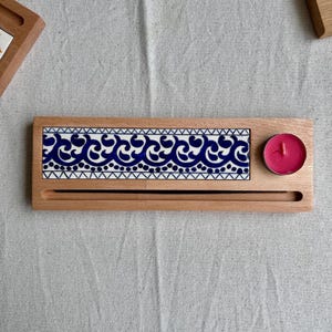 May include: A rectangular wooden tray featuring an inlaid ceramic tile with a blue and white wave pattern. The tray includes a long, narrow horizontal slot for incense sticks and a circular cutout holding a small, round, bright pink tea light candle.