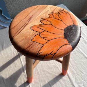 Puede incluir: Un taburete redondo de madera con un diseño de girasol naranja pintado a mano en el asiento. El taburete tiene tres patas de madera clara, cada una con un detalle pintado en naranja brillante en la parte inferior, manteniendo el estilo.