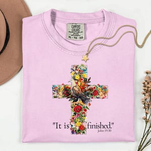 May include: A light pink Comfort Colors t-shirt with a floral cross design and a dove. The cross is adorned with colorful flowers and a dove. The text "It is finished." and John 19:30 are printed below the cross. A gold necklace with a star pendant is on the shirt.