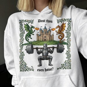 Pu&ograve; includere: Felpa con cappuccio bianca con grafica a tema medievale. Il design presenta un cavaliere che solleva pesi, affiancato da draghi, con un castello sullo sfondo. Il testo recita "Dost thou even hoist?" in un font medievale.